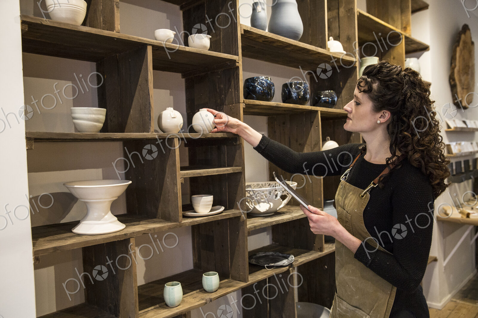 Woman with curly brown hair wearing apron standing in her pottery shop, arranging…