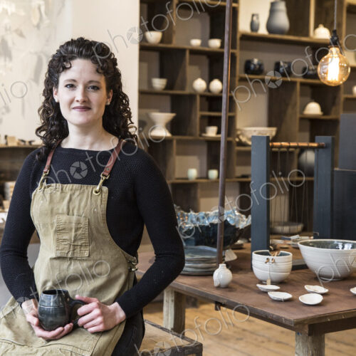 Woman with curly brown hair wearing apron standing in her pottery shop, holding mug,… Woman with curly brown hair wearing apron standing in her pottery shop, holding mug,…