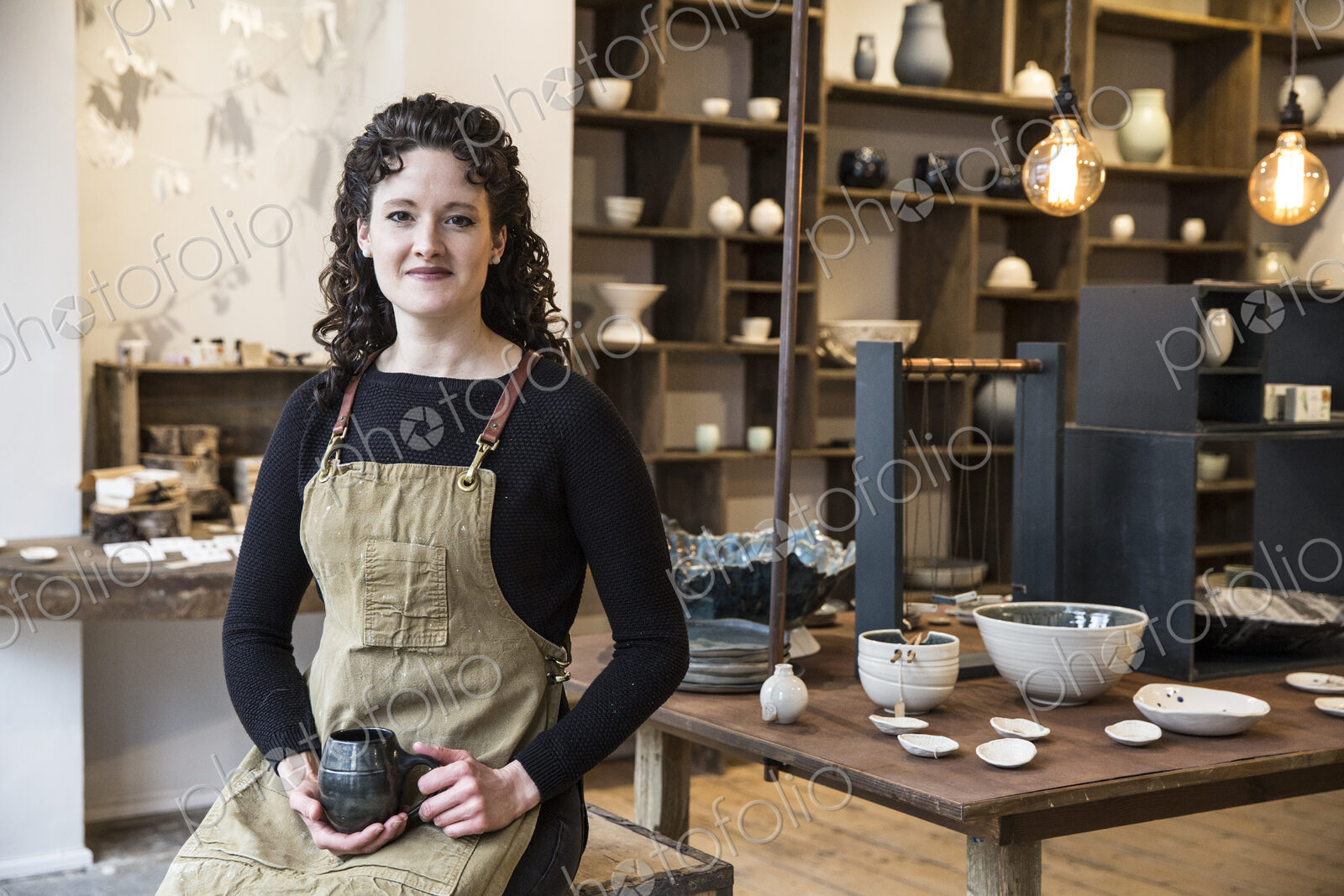 Woman with curly brown hair wearing apron standing in her pottery shop, holding mug,…