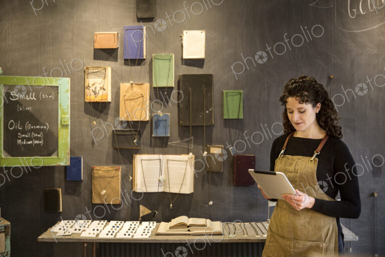 Woman with curly brown hair wearing apron standing in her pottery shop, looking at…