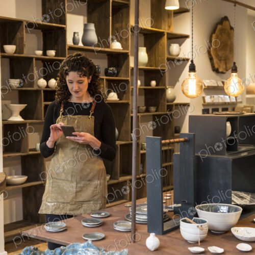 Woman with curly brown hair wearing apron standing in her pottery shop, looking at… Woman with curly brown hair wearing apron standing in her pottery shop, looking at…