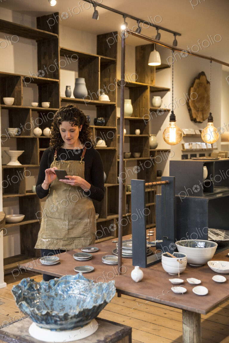 Woman with curly brown hair wearing apron standing in her pottery shop, looking at…