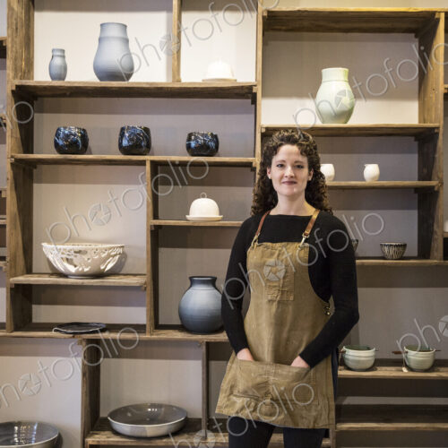 Woman with curly brown hair wearing apron standing in her pottery shop, smiling at camera. Woman with curly brown hair wearing apron standing in her pottery shop, smiling at camera.