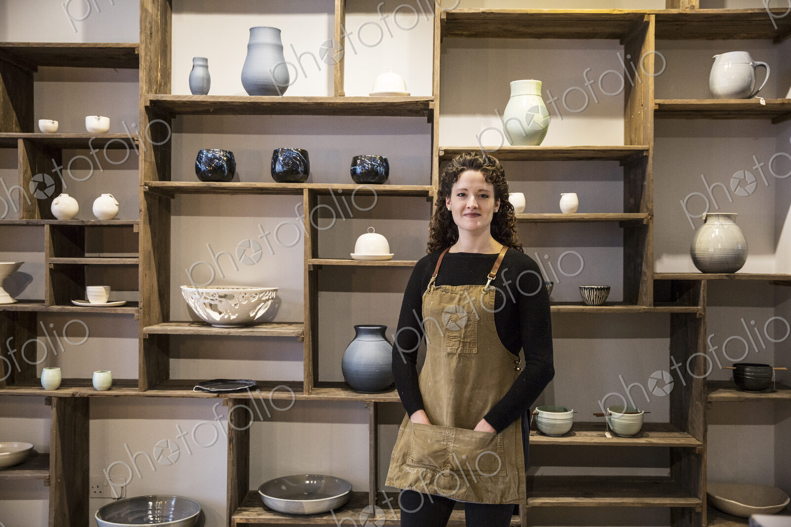Woman with curly brown hair wearing apron standing in her pottery shop, smiling at camera.