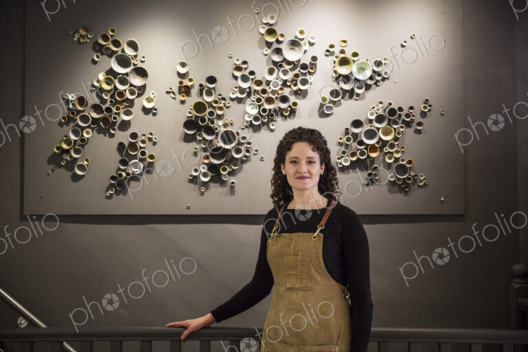 Woman with curly brown hair wearing apron standing in her pottery shop, smiling at camera.