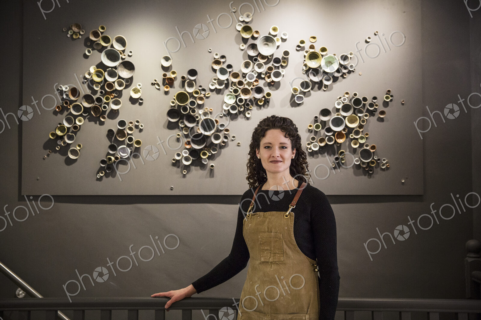 Woman with curly brown hair wearing apron standing in her pottery shop, smiling at camera.