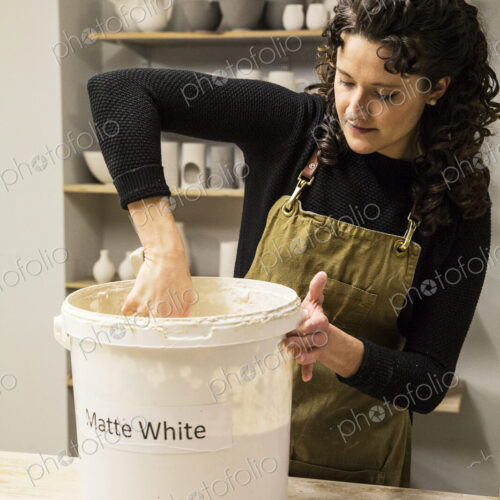 Woman with curly brown hair wearing apron standing in pottery workshop, dipping vase… Woman with curly brown hair wearing apron standing in pottery workshop, dipping vase…