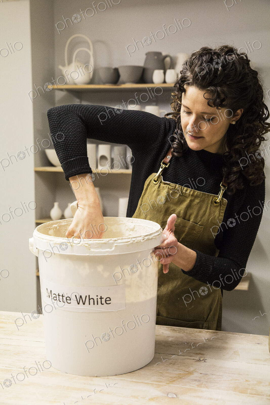 Woman with curly brown hair wearing apron standing in pottery workshop, dipping vase…