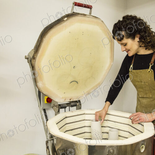 Woman with curly brown hair wearing apron standing in pottery workshop, placing vase… Woman with curly brown hair wearing apron standing in pottery workshop, placing vase…