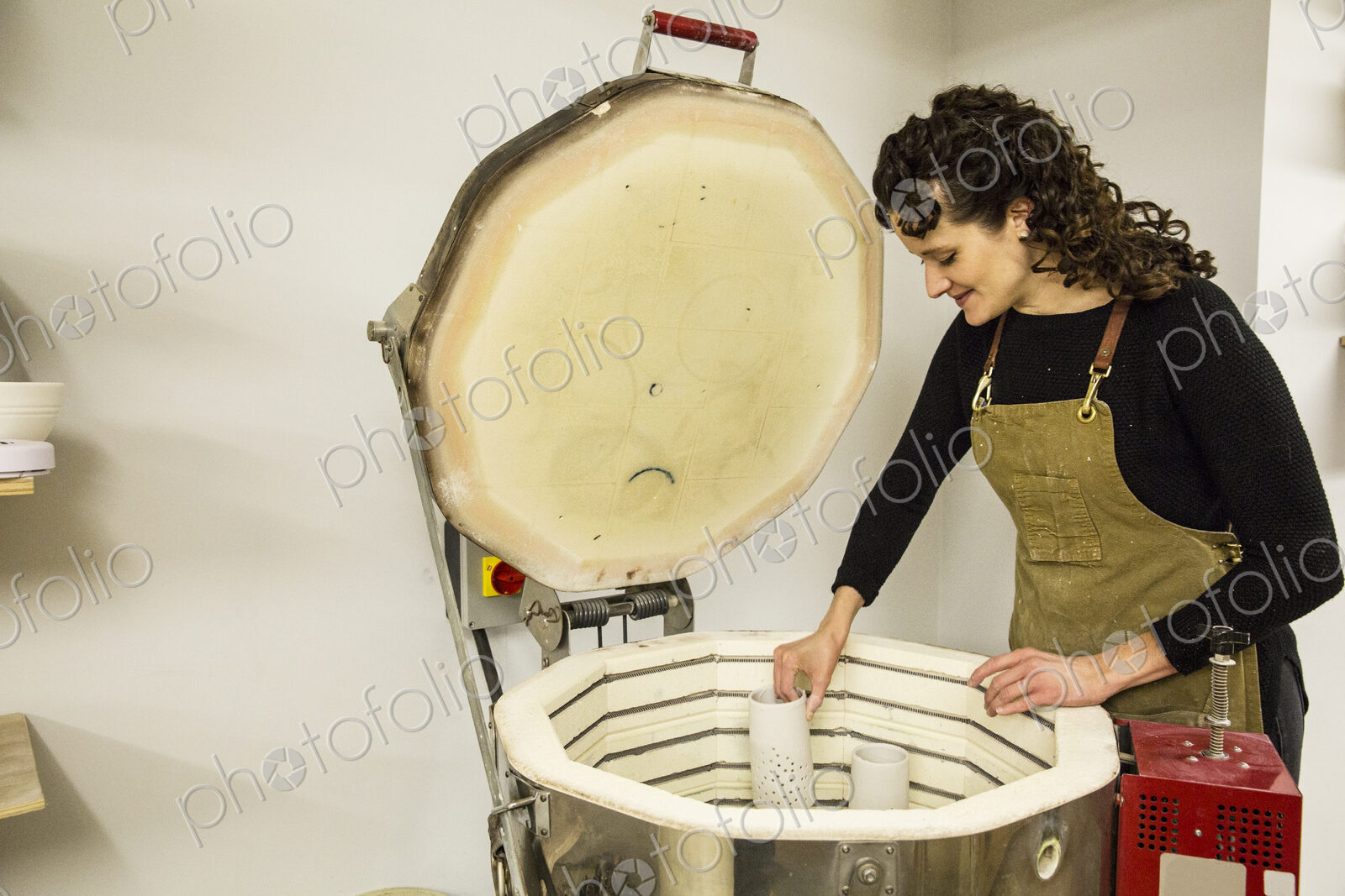 Woman with curly brown hair wearing apron standing in pottery workshop, placing vase…