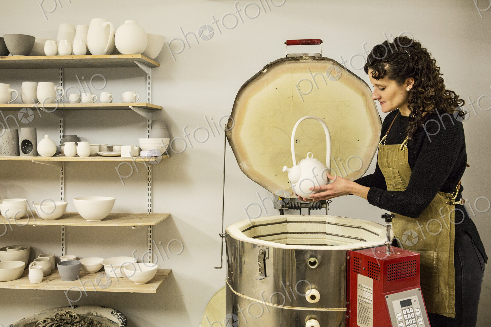 Woman with curly brown hair wearing apron standing in pottery workshop, placing vase…