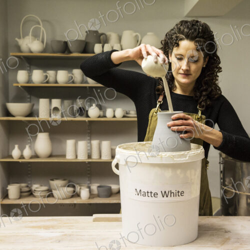 Woman with curly brown hair wearing apron standing in pottery workshop, pouring white… Woman with curly brown hair wearing apron standing in pottery workshop, pouring white…