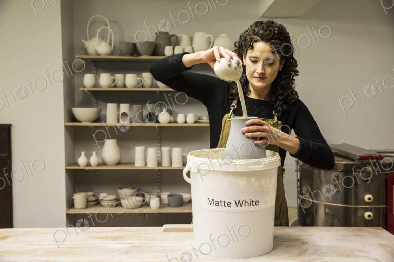 Woman with curly brown hair wearing apron standing in pottery workshop, pouring white…