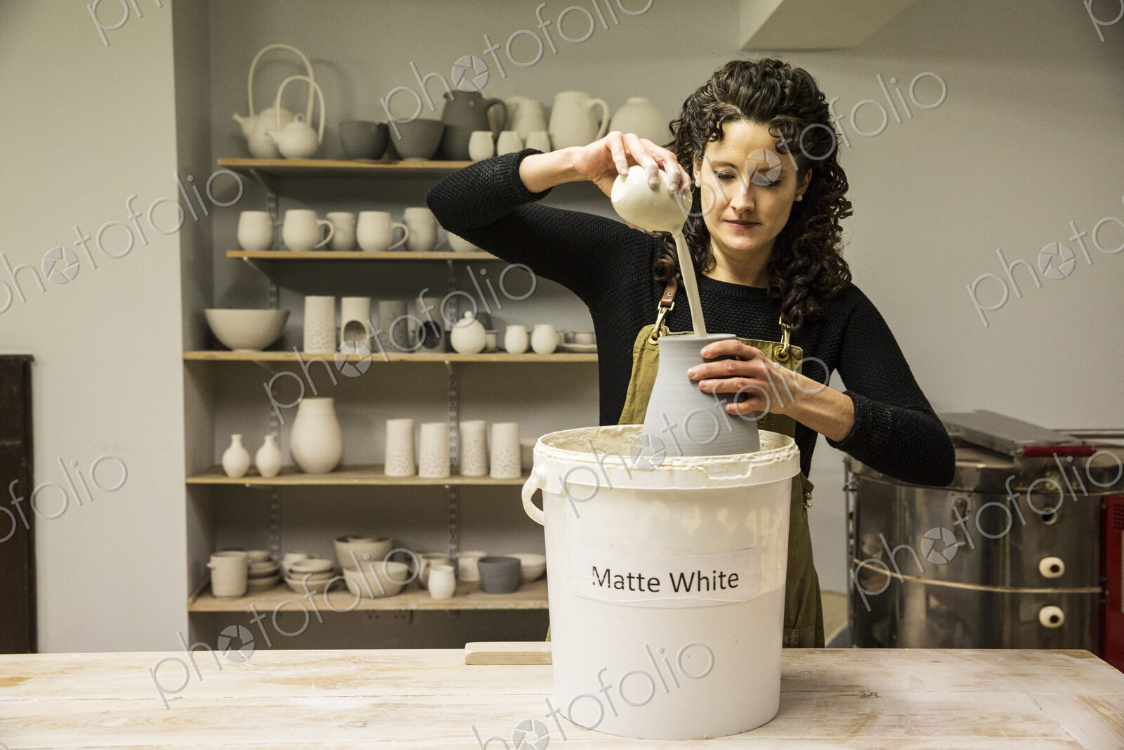 Woman with curly brown hair wearing apron standing in pottery workshop, pouring white…