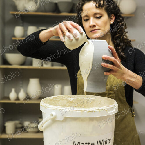 Woman with curly brown hair wearing apron standing in pottery workshop, pouring white… Woman with curly brown hair wearing apron standing in pottery workshop, pouring white…