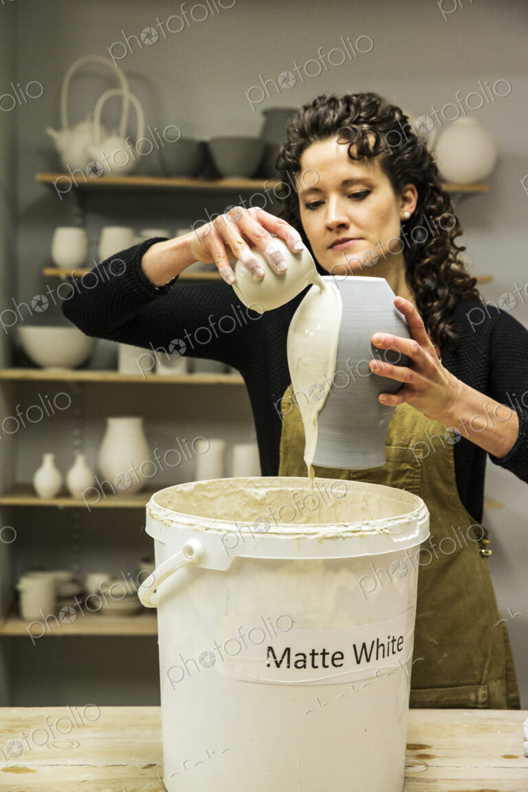 Woman with curly brown hair wearing apron standing in pottery workshop, pouring white…