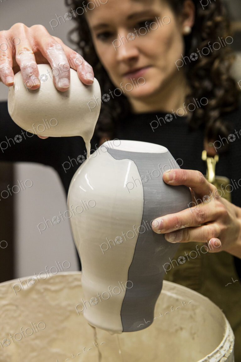 Woman with curly brown hair wearing apron standing in pottery workshop, pouring white…