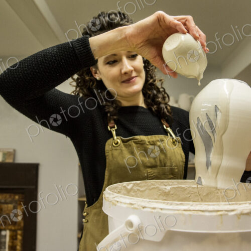 Woman with curly brown hair wearing apron standing in pottery workshop, pouring white… Woman with curly brown hair wearing apron standing in pottery workshop, pouring white…