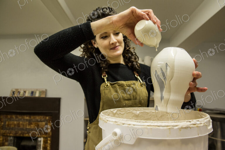 Woman with curly brown hair wearing apron standing in pottery workshop, pouring white…