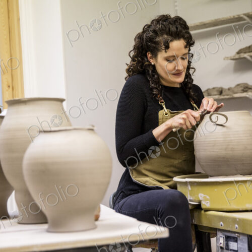 Woman with curly brown hair wearing apron working on spherical clay vase on pottery wheel. Woman with curly brown hair wearing apron working on spherical clay vase on pottery wheel.