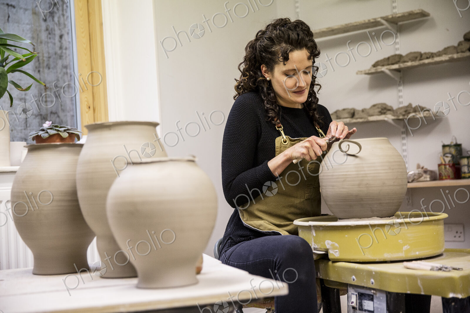 Woman with curly brown hair wearing apron working on spherical clay vase on pottery wheel.
