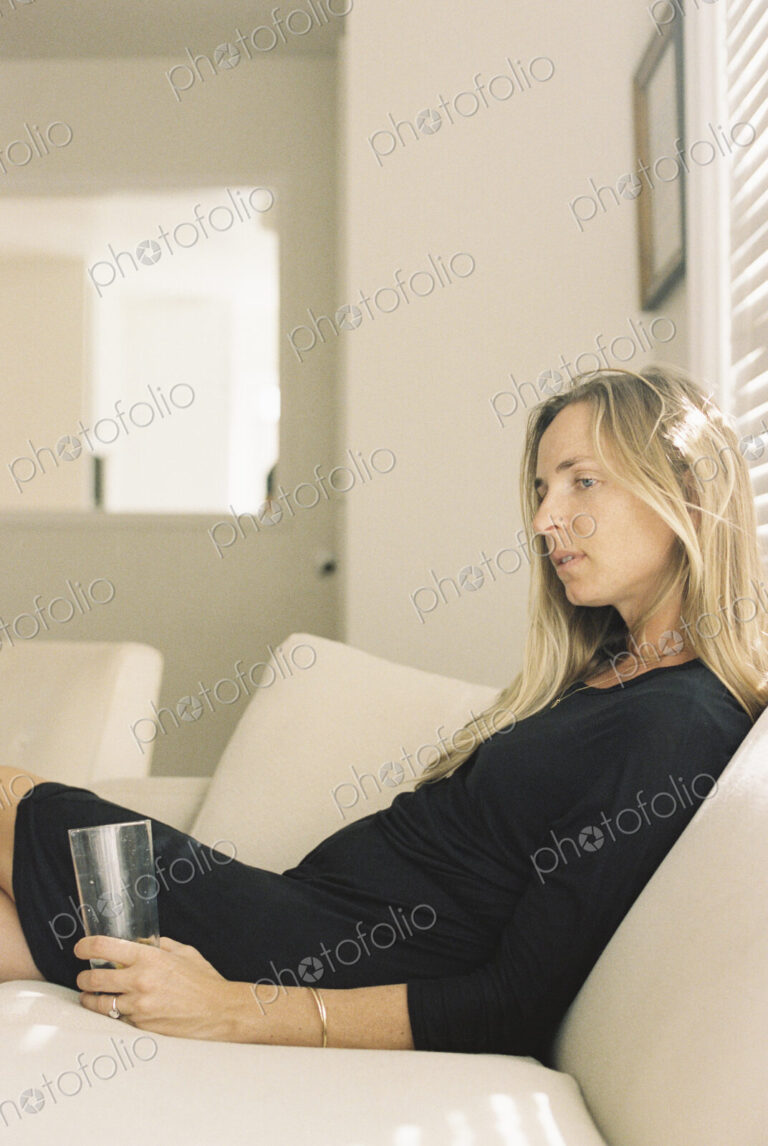Woman with long blond hair wearing a black dress, sitting on a sofa, holding a glass.