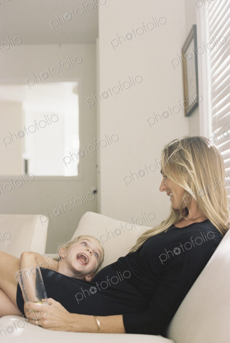 Woman with long blond hair wearing a black dress, sitting on a sofa, holding a glass,…