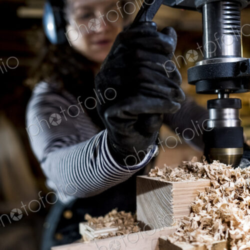 Woman with long brown hair wearing dungarees, safety glasses and ear protectors…