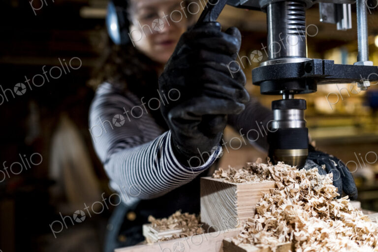 Woman with long brown hair wearing dungarees, safety glasses and ear protectors…
