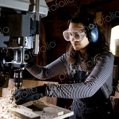 Woman with long brown hair wearing dungarees, safety glasses and ear protectors…