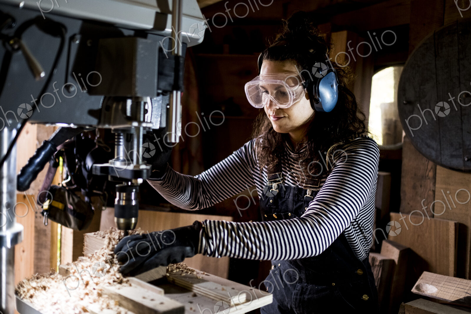 Woman with long brown hair wearing dungarees, safety glasses and ear protectors…