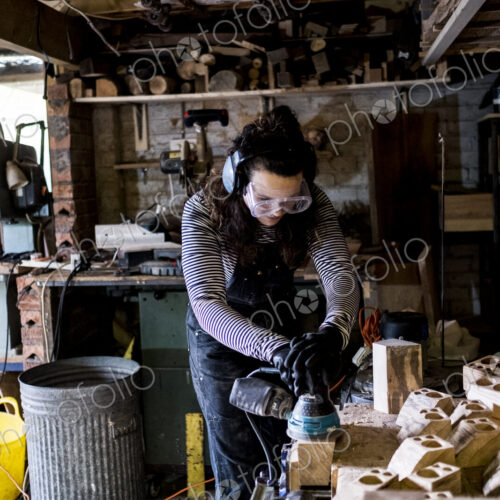 Woman with long brown hair wearing dungarees, safety glasses and ear protectors…