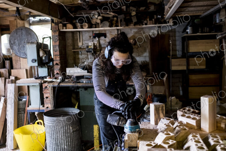 Woman with long brown hair wearing dungarees, safety glasses and ear protectors…