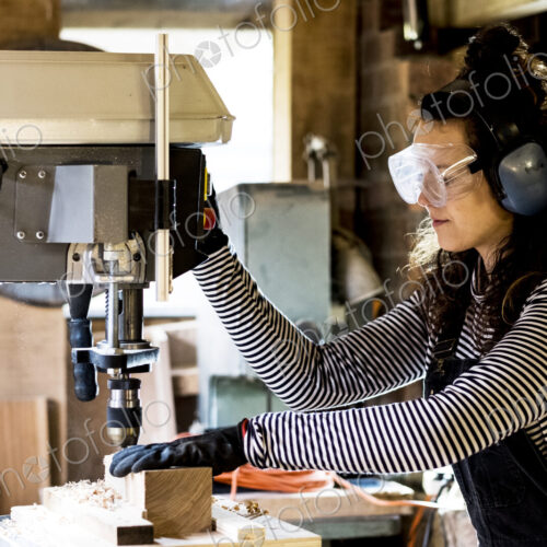 Woman with long brown hair wearing dungarees, safety glasses and ear protectors…