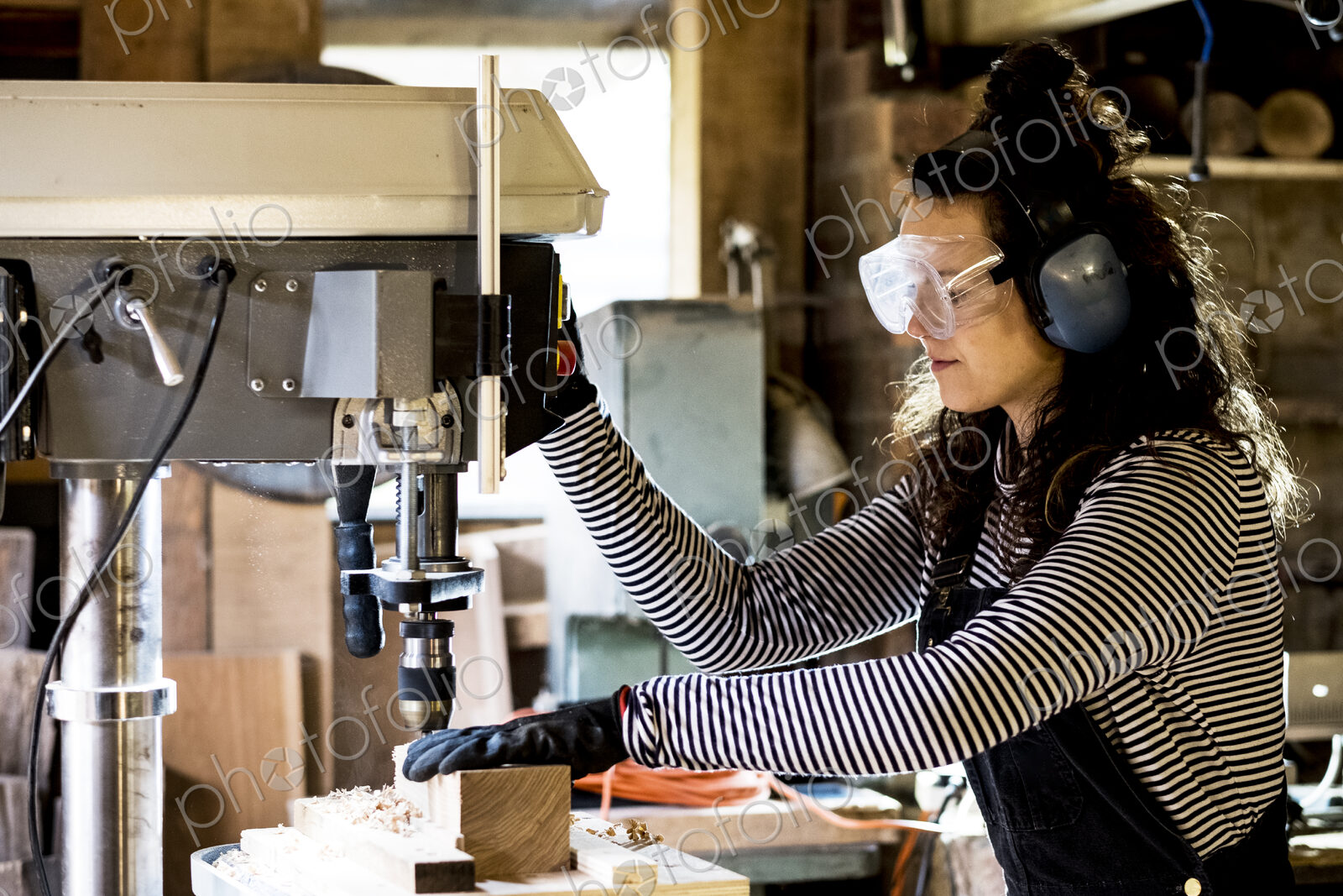 Woman with long brown hair wearing dungarees, safety glasses and ear protectors…