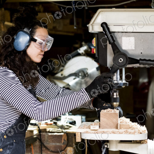 Woman with long brown hair wearing dungarees, safety glasses and ear protectors…