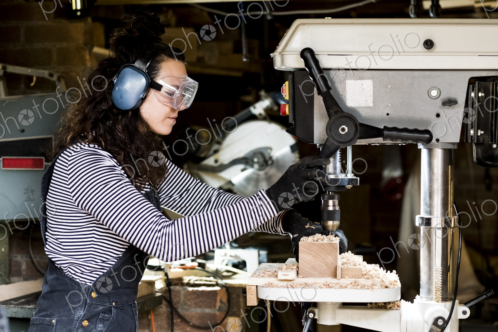 Woman with long brown hair wearing dungarees, safety glasses and ear protectors…