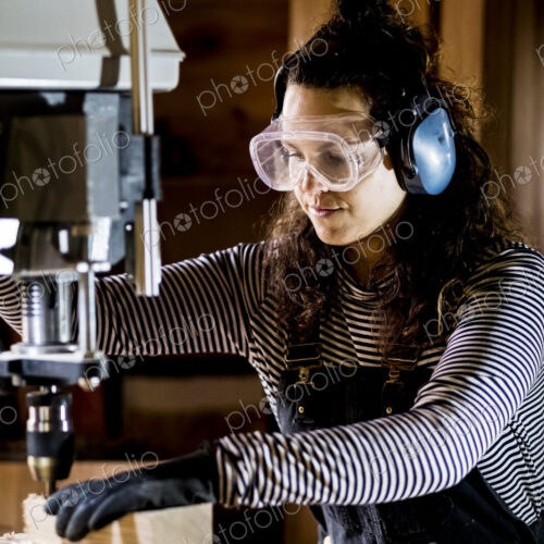 Woman with long brown hair wearing dungarees, safety glasses and ear protectors…