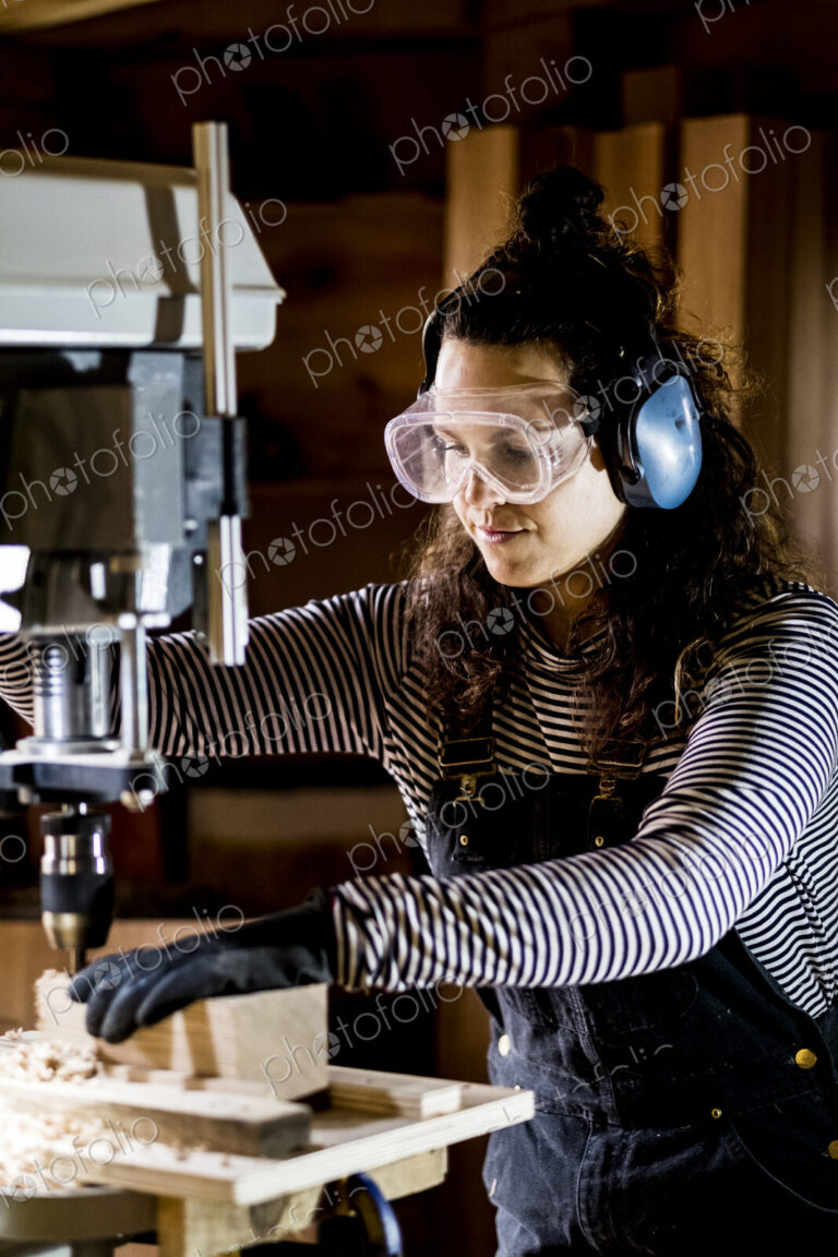 Woman with long brown hair wearing dungarees, safety glasses and ear protectors…