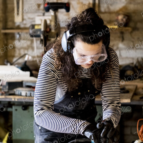 Woman with long brown hair wearing dungarees, safety glasses and ear protectors…