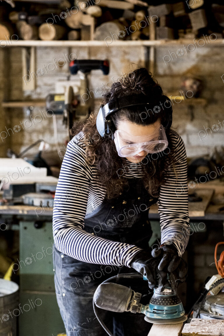 Woman with long brown hair wearing dungarees, safety glasses and ear protectors…