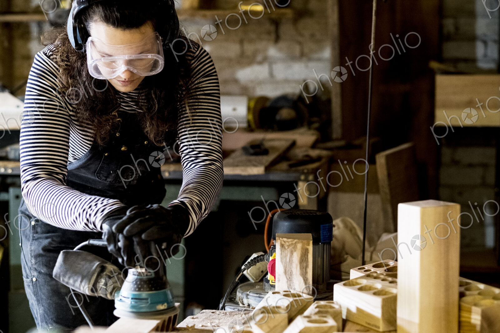 Woman with long brown hair wearing dungarees, safety glasses and ear protectors…