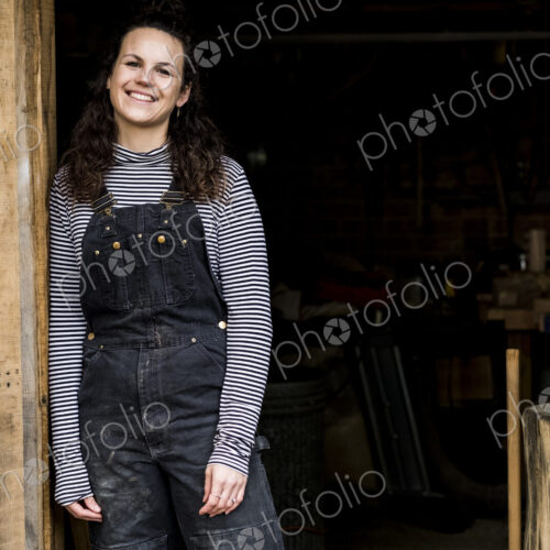 Woman with long brown hair wearing dungarees standing at entrance to wood workshop,… Woman with long brown hair wearing dungarees standing at entrance to wood workshop,…