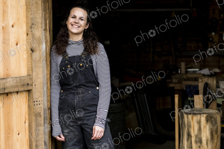 Woman with long brown hair wearing dungarees standing at entrance to wood workshop,…