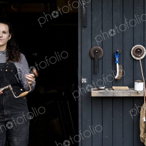 Woman with long brown hair wearing dungarees standing at entrance to wood workshop,… Woman with long brown hair wearing dungarees standing at entrance to wood workshop,…