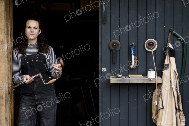 Woman with long brown hair wearing dungarees standing at entrance to wood workshop,…
