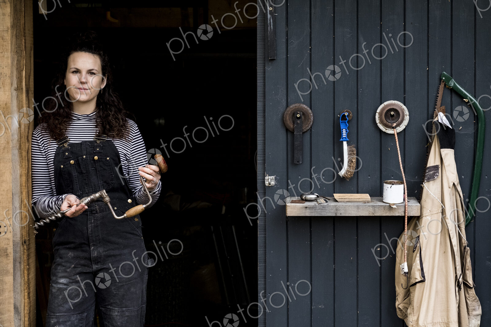 Woman with long brown hair wearing dungarees standing at entrance to wood workshop,…