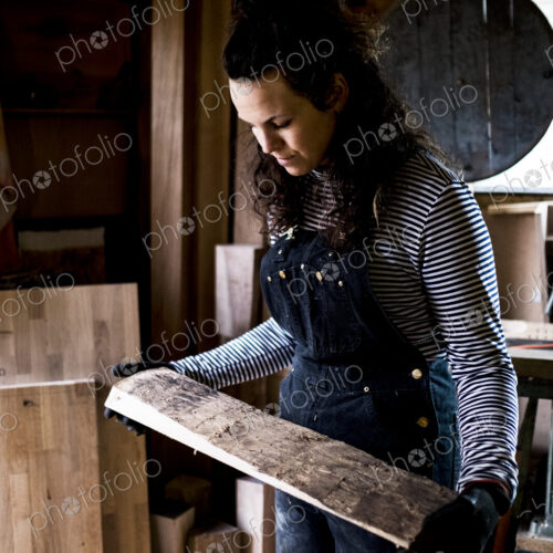 Woman with long brown hair wearing dungarees standing in wood workshop, holding piece… Woman with long brown hair wearing dungarees standing in wood workshop, holding piece…