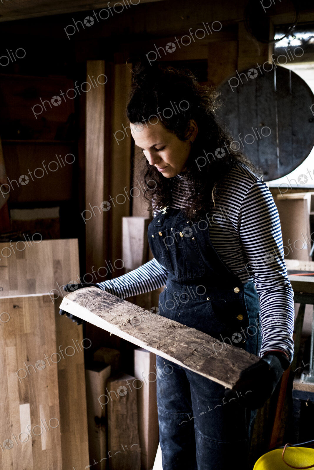 Woman with long brown hair wearing dungarees standing in wood workshop, holding piece…