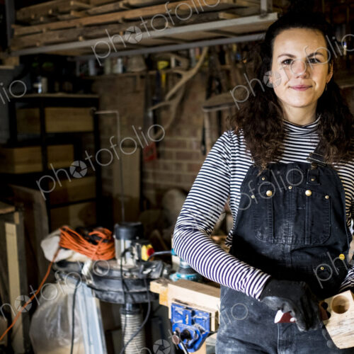 Woman with long brown hair wearing dungarees standing in wood workshop, smiling at camera. Woman with long brown hair wearing dungarees standing in wood workshop, smiling at camera.